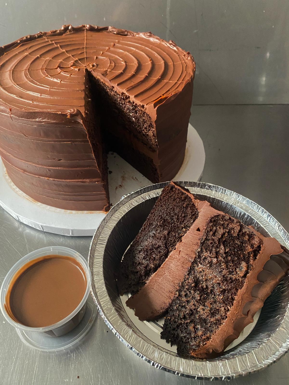 A chocolate layer cake with Belgian milk chocolate ganache frosting, served on a plate with a cup of coffee. A pot of warm Belgian milk chocolate sauce is also visible.